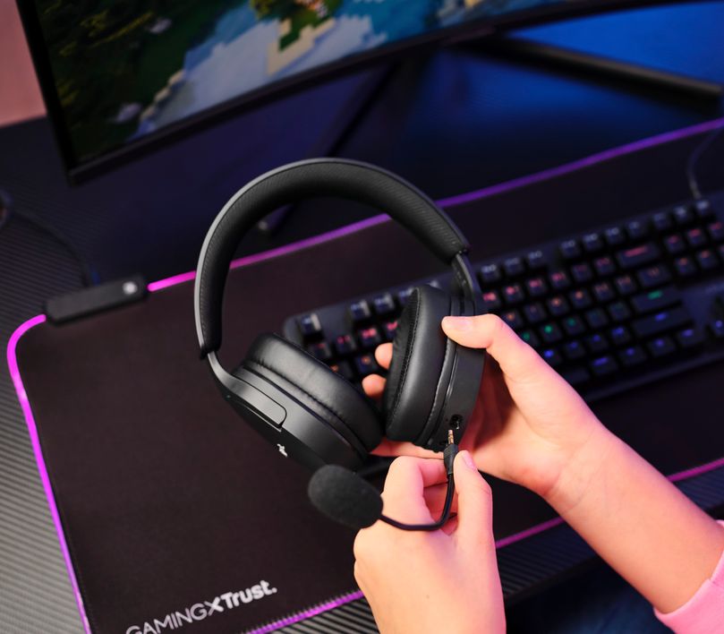 Manos sosteniendo unos Auriculares Gaming Inalámbricos Trust GXT 491 Fayzo, de color negro sobre un escritorio con teclado y mousepad iluminados por luces LED.