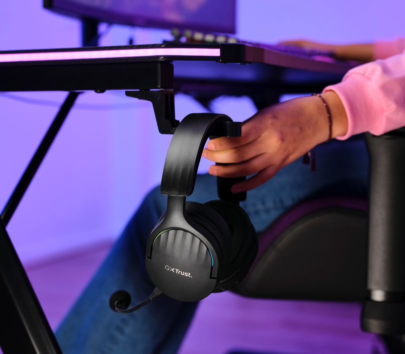Auriculares Gaming Inalámbricos Trust GXT 491 Fayzo, de color Negro colgando de un soporte para auriculares adherido a una mesa, con una persona al fondo tocandolo, iluminado por luces azules y violetas.