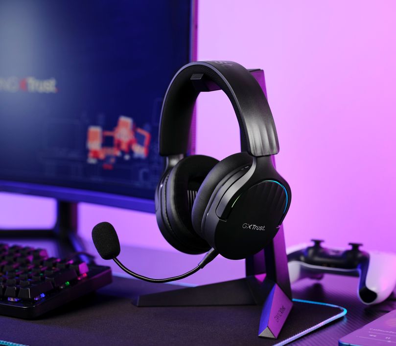Auriculares Gaming Inalámbricos Trust GXT 491 Fayzo, de color Negro, apoyados en un soporte, frente a una pantalla de computadora y un teclado en un fondo iluminado con luces violetas y azules.