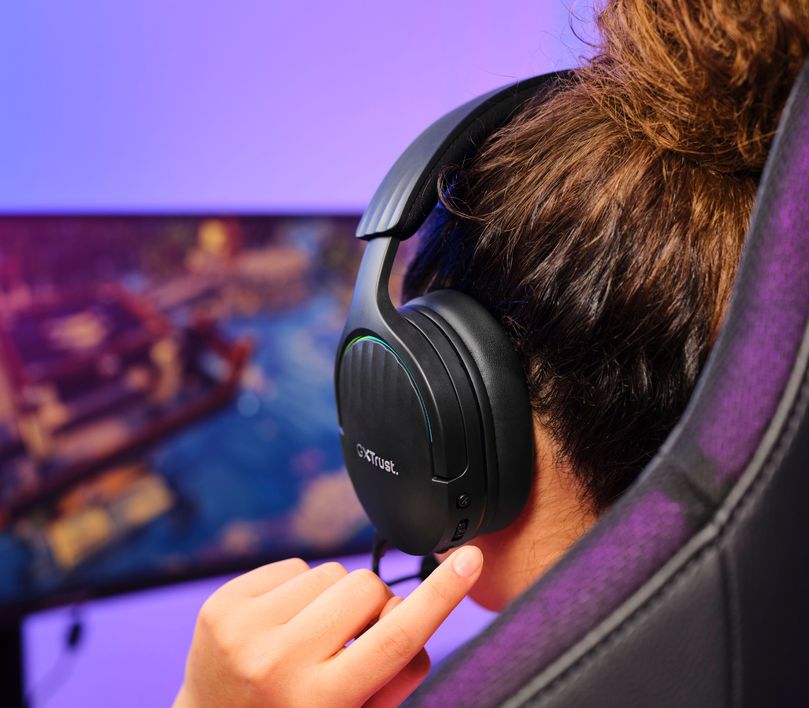 Persona con Auriculares Gaming Inalámbricos Trust GXT 491 Fayzo, de color Negro ajustando el volumen en un dial lateral, sentada frente a una computadora con pantalla mostrando gráficos coloridos de un videojuego.