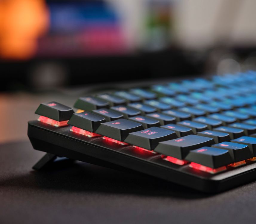 Vista del Teclado mecánico prémium Trust GXT 866 Torix, con iluminación LED de color rojo. sobre una mesa, enfoque selectivo en las teclas del primer plano y fondo desenfocado con pantalla de computadora.