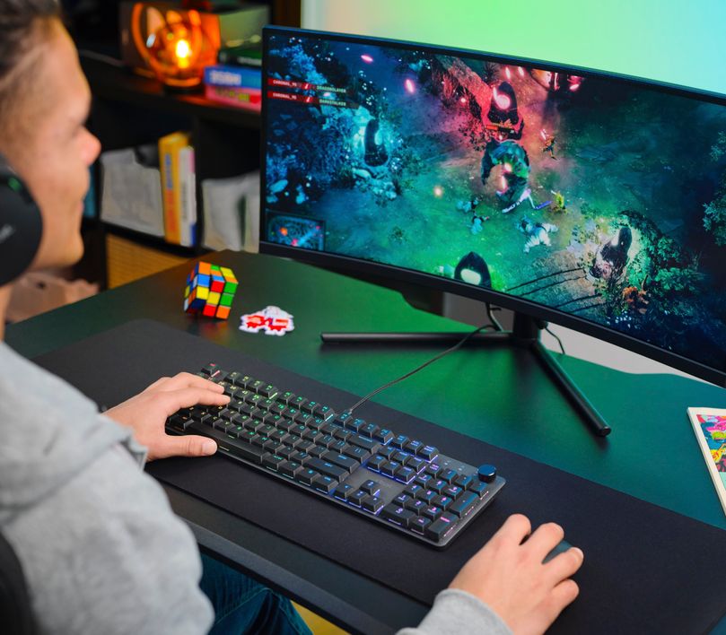 Persona utilizando un Teclado mecánico prémium Trust GXT 866 Torix frente a un monitor curvo con una imagen colorida de un videojuego.
