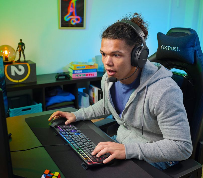 Persona con auriculares jugando en una computadora con un Teclado mecánico prémium Trust GXT 866 Torix, en una habitación iluminada con luces de colores y decorada con figuras de acción y un cubo Rubik.