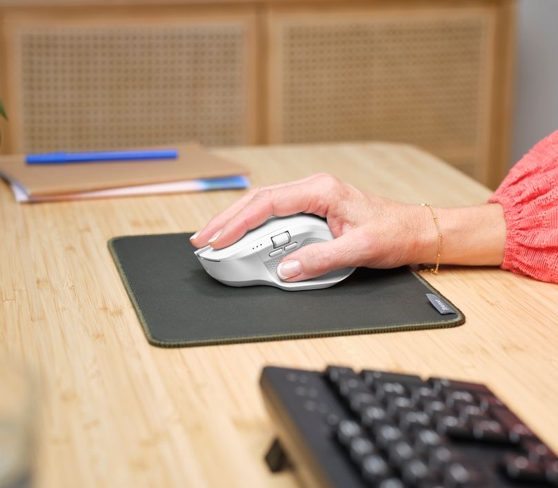 Una mano usando un Mouse inalámbrico multidispositivo Trust Ozaa+ de color blanco sobre una alfombrilla gris en un escritorio de madera clara. Se puede ver también un teclado negro y unos cuadernos con un bolígrafo azul encima.