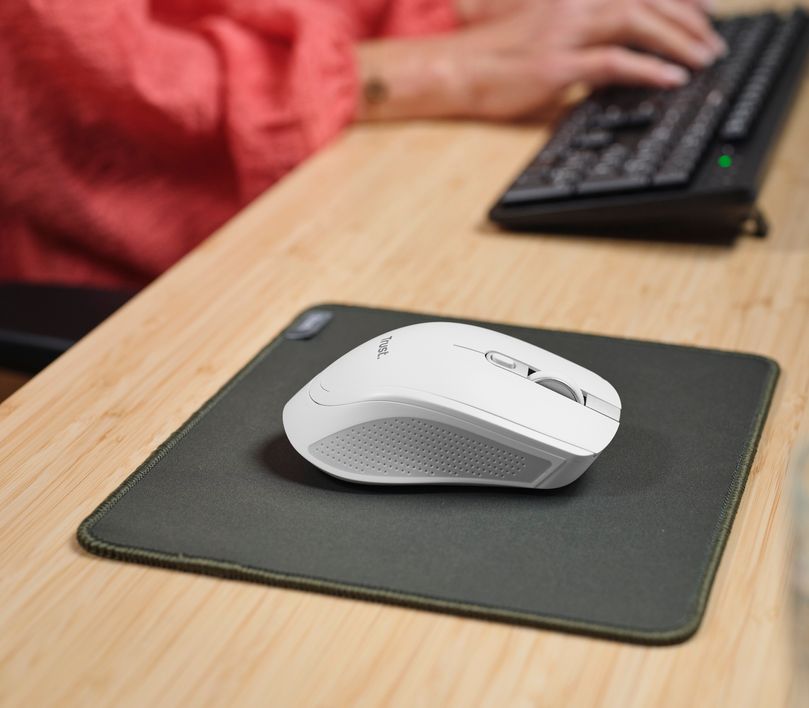 Un Mouse inalámbrico multidispositivo Trust Ozaa+ de color blanco sobre una alfombrilla gris en un escritorio de madera clara. Al fondo, una persona está escribiendo en un teclado.