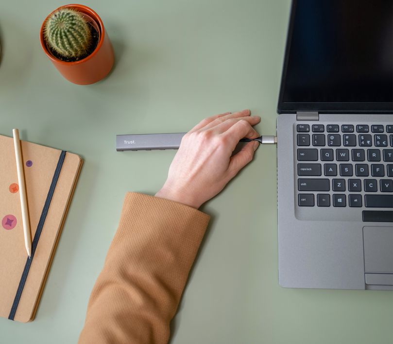 Una mano conecta un Hub USB-C de 4 puertos USB-A 3.2 Gen1 Trust Halyx a una laptop sobre un escritorio con cuadernos, lápices y una planta suculenta.