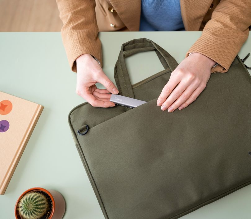 Una persona coloca un Hub USB-C de 4 puertos USB-A 3.2 Gen1 Trust Halyx en el bolsillo frontal de un maletín verde oliva sobre una mesa, junto a un libro con tapa naranja decorada con corazones y una planta suculenta en una maceta.