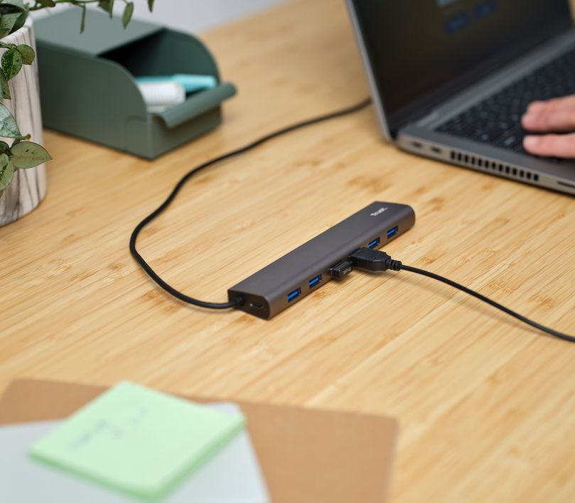 Un Hub USB 3.2 Gen1 de 7 puertos Trust Halyx conectado a una laptop sobre un escritorio de madera, con objetos de oficina como un organizador verde, una planta y notas adhesivas alrededor.