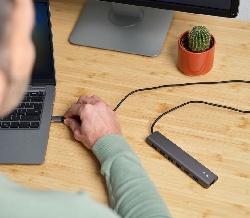 Persona conectando un Hub USB 3.2 Gen1 de 7 puertos Trust Halyx a una laptop en una oficina en casa, con un monitor adicional y un cactus en maceta naranja sobre la mesa.