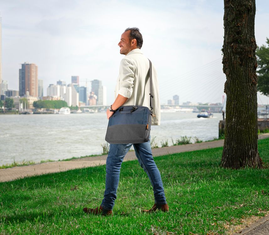 Una persona de pie en un parque junto a un río, con una ciudad moderna y un puente al fondo. Lleva una chaqueta clara, jeans y zapatos marrones, y sostiene un Maletín para Laptop de 16 pulgadas Trust Lisboa de color azul