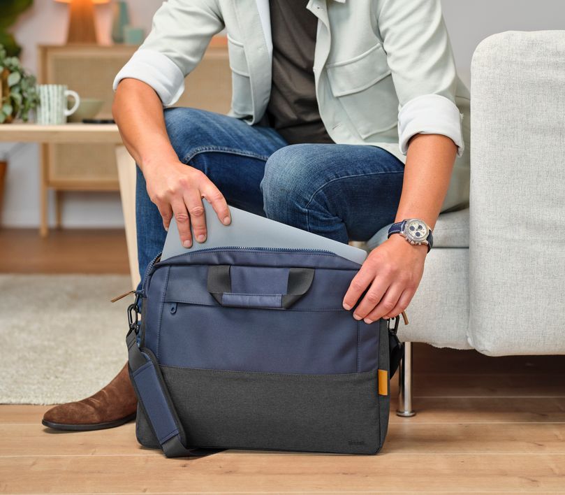 Una persona está sentada en un sofá, sacando una laptop de un Maletín para Laptop de 16 pulgadas Trust Lisboa de color azul. La persona lleva una camisa clara, jeans y un reloj en la muñeca izquierda. En el fondo, se puede ver una mesa con una taza y algunos objetos decorativos.
