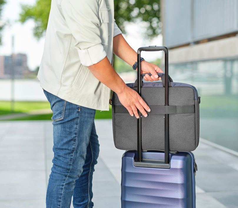 Una persona con una chaqueta clara y jeans está colocando un Maletín para Laptop de 16 pulgadas Trust Lisboa de color azul sobre el asa extendida de una maleta de viaje azul. La escena se desarrolla en un entorno exterior, posiblemente cerca de un edificio moderno y un área verde.