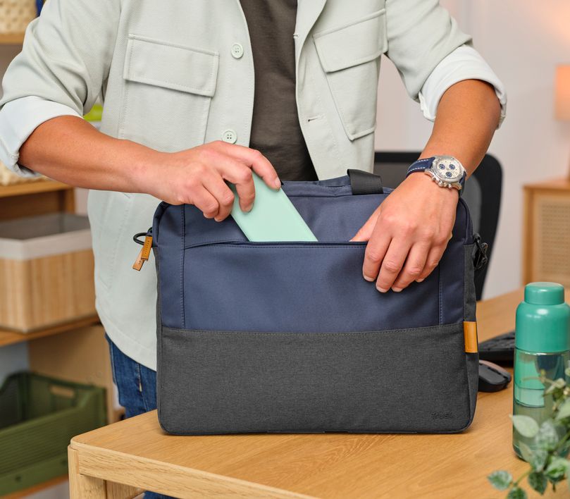 Una persona está guardando un objeto rectangular verde en el bolsillo frontal de un Maletín para Laptop de 16 pulgadas Trust Lisboa de color azul. La persona lleva una chaqueta clara y un reloj plateado en la muñeca izquierda. En la mesa de madera hay una botella verde y algunos objetos más en el fondo.