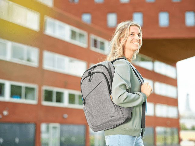 Mujer joven de cabello rubio, con una expresión alegre y sonriente, de pie al aire libre llevando la mochila Trust Avana de color gris sobre sus hombros.