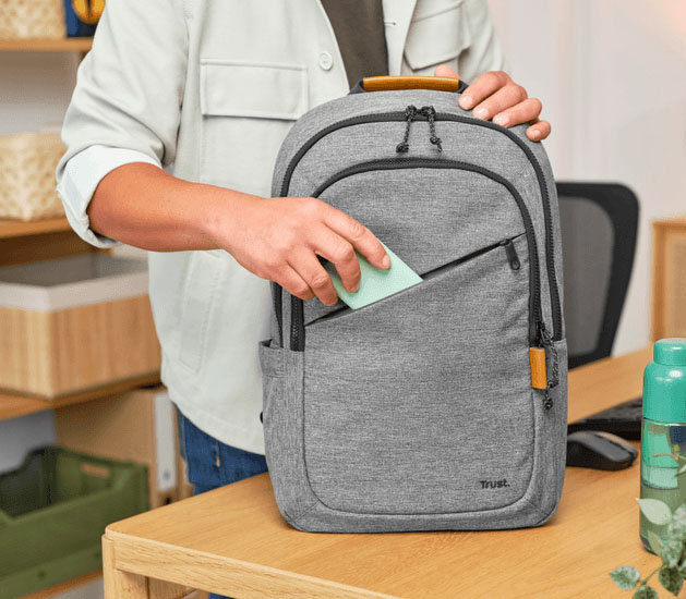Manos insertando un objeto verde claro en el bolsillo frontal con cremallera de la mochila Trust Avana gris, sobre un escritorio de madera en un ambiente de oficina.