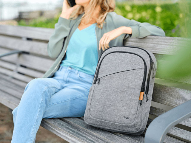 La mochila Trust Avana de color gris descansa en primer plano sobre un banco de madera al aire libre, junto a una mujer sentada que habla por teléfono.