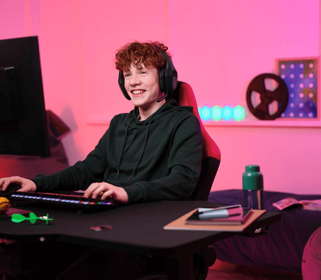 Adolescente con Auriculares Gaming Livianos Trust GXT 415 Zirox de color negro, jugando en una PC, sentado en una silla ergonómica frente a un escritorio con teclado RGB, botella de agua y útiles, en una habitación iluminada con luces LED rosas.