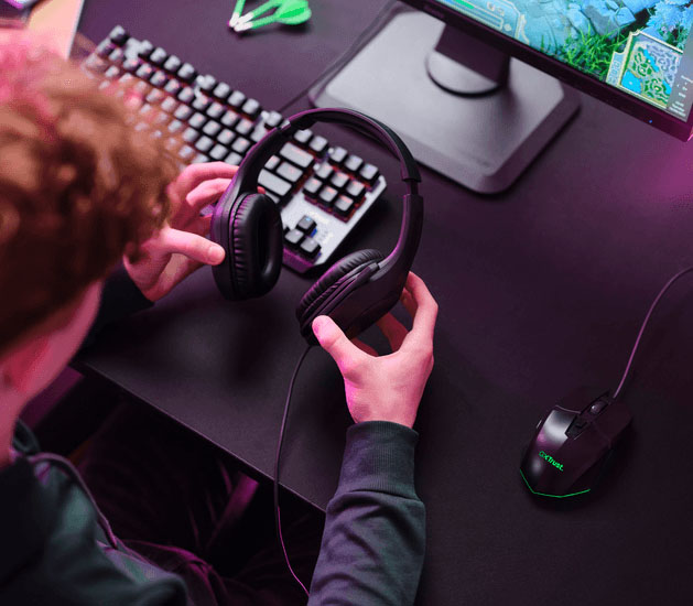 Persona joven sosteniendo unos Auriculares Gaming Livianos Trust GXT 415 Zirox de color negro, frente a una computadora, con teclado mecánico, mouse gamer y monitor encendido, en un entorno iluminado con luces LED de colores.