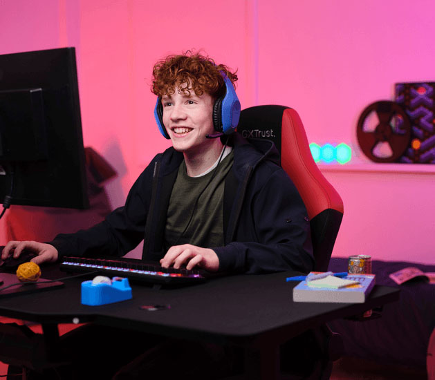 Adolescente con Auriculares Gaming Livianos Trust GXT 415B Zirox de color azul, jugando en una PC, sentado en una silla ergonómica frente a un escritorio con teclado RGB, botella de agua y útiles, en una habitación iluminada con luces LED rosas.