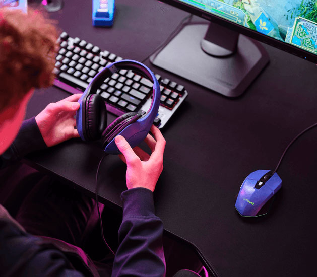 Persona joven sosteniendo unos Auriculares Gaming Livianos Trust GXT 415B Zirox de color azul, frente a una computadora, con teclado mecánico, mouse gamer y monitor encendido, en un entorno iluminado con luces LED de colores.