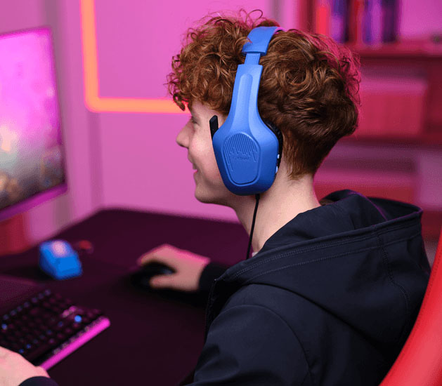 Adolescente con Auriculares Gaming Livianos Trust GXT 415B Zirox de color azul, jugando en una PC, sonriendo mientras usa el mouse, frente a un monitor con iluminación LED rosa y teclado retroiluminado.