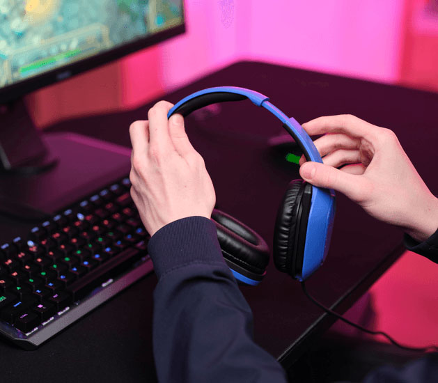 Primer plano de manos ajustando unos Auriculares Gaming Livianos Trust GXT 415B Zirox de color azul, frente a un monitor con videojuego, teclado mecánico retroiluminado y luces LED rosas de fondo.