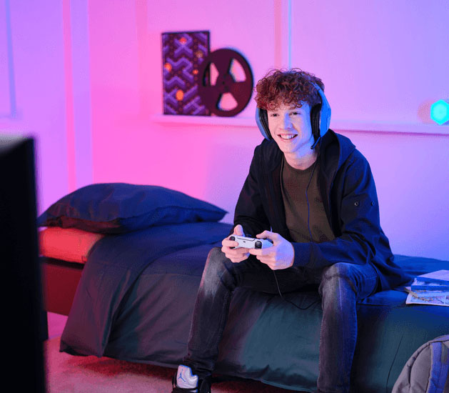 Adolescente sentado en la cama jugando con un joystick y Auriculares Gaming Livianos Trust GXT 415B Zirox de color azul, disfrutando de un videojuego frente a una pantalla, en una habitación con luces LED en tonos rosa y azul.