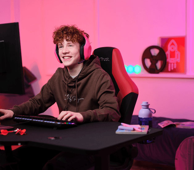 Adolescente con Auriculares Gaming Livianos Trust GXT 415P Zirox de color rosa, jugando en una PC, sentado en una silla ergonómica frente a un escritorio con teclado RGB, botella de agua y útiles, en una habitación iluminada con luces LED rosas.
