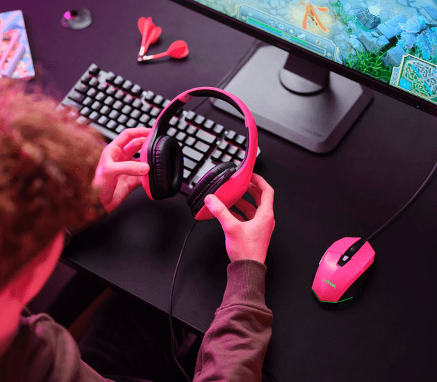 Persona joven sosteniendo unos Auriculares Gaming Livianos Trust GXT 415P Zirox de color rosa, frente a una computadora, con teclado mecánico, mouse gamer y monitor encendido, en un entorno iluminado con luces LED de colores.
