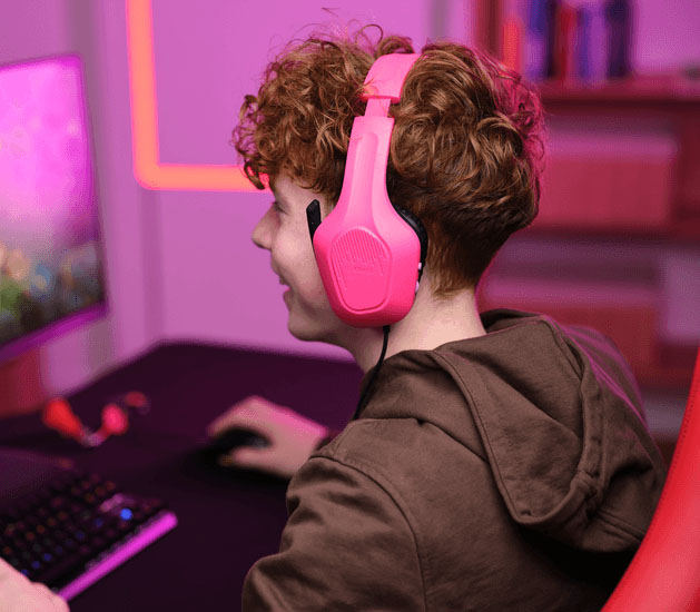 Adolescente con Auriculares Gaming Livianos Trust GXT 415P Zirox de color rosa, jugando en una PC, sonriendo mientras usa el mouse, frente a un monitor con iluminación LED rosa y teclado retroiluminado.