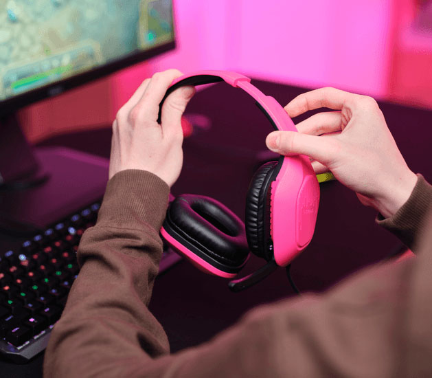 Primer plano de manos ajustando unos Auriculares Gaming Livianos Trust GXT 415P Zirox de color rosa, frente a un monitor con videojuego, teclado mecánico retroiluminado y luces LED rosas de fondo.