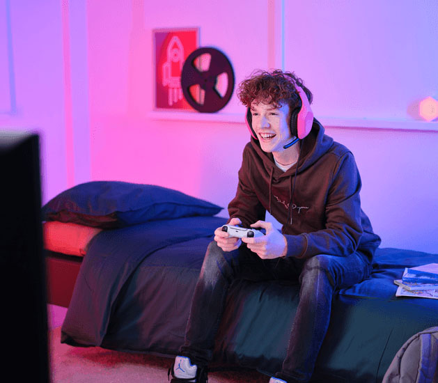 Adolescente sentado en la cama jugando con un joystick y Auriculares Gaming Livianos Trust GXT 415P Zirox de color rosa, disfrutando de un videojuego frente a una pantalla, en una habitación con luces LED en tonos rosa y azul.