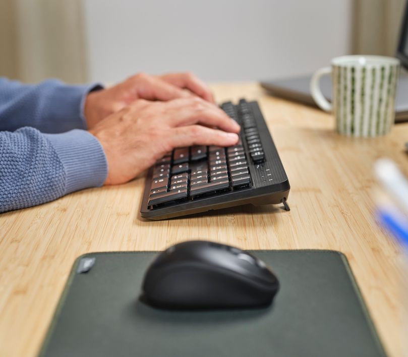 Mano sobre un teclado de computadora con una taza y un mouse al lado en un escritorio de madera.