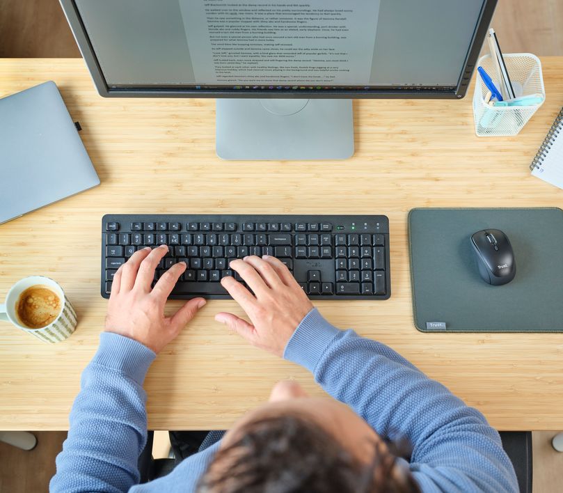 Vista aérea de una persona utilizando un Combo de teclado y mouse inalámbricos y silenciosos Trust Ody II en un escritorio con una computadora, taza de café y otros artículos de oficina.