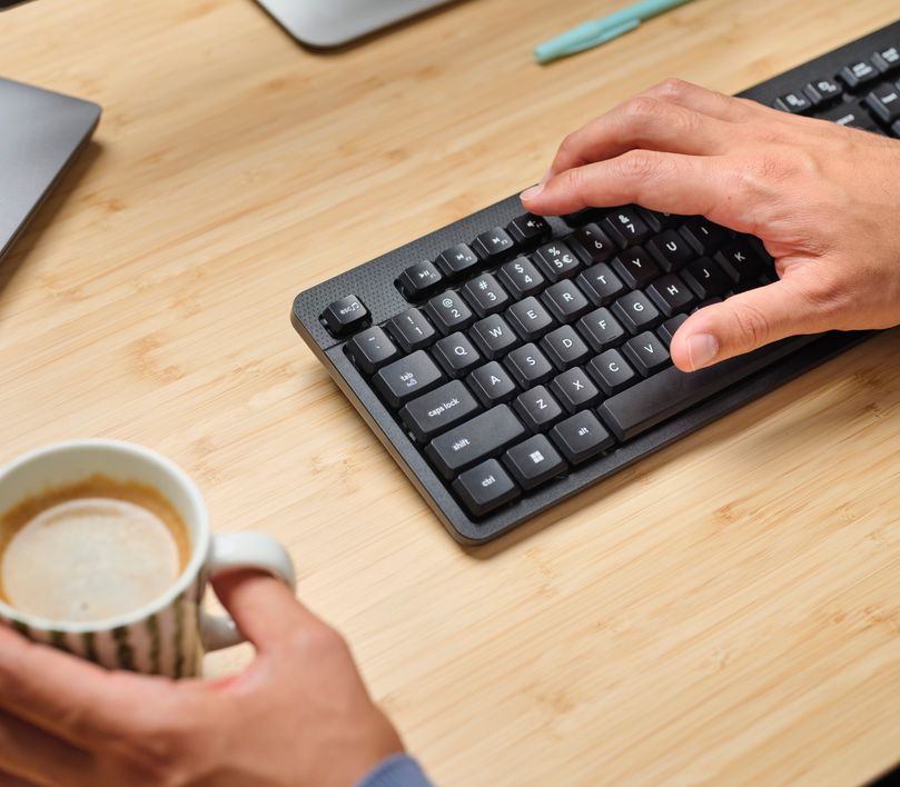 Mano izquierda sosteniendo una taza de café cerca del borde de una mesa de madera, mientras la mano derecha pulsa una tecla de acceso rápido en un teclado negro.