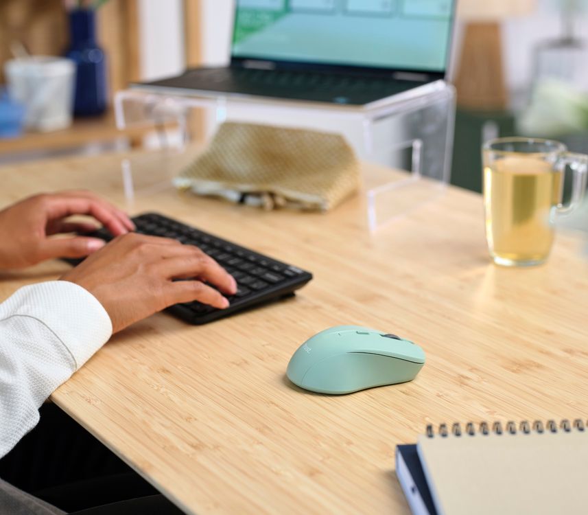 Imagen de un escritorio con una computadora portátil con gráficos en la pantalla, una taza de té, y manos tecleando en un teclado inalámbrico. Al lado del teclado hay un Mouse inalámbrico silencioso Trust Mydo de color verde