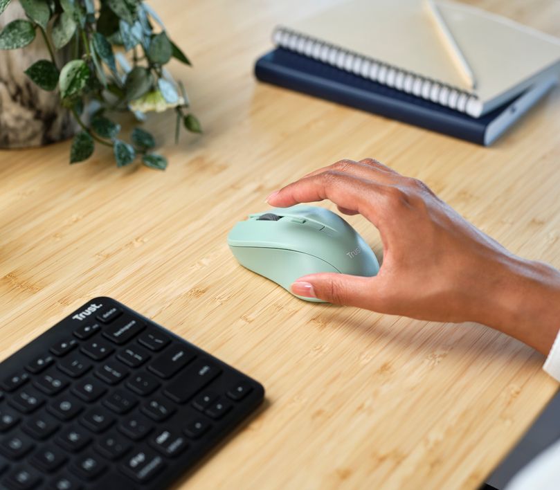 Una mano sobre un Mouse inalámbrico silencioso Trust Mydo de color verde al lado de un teclado y una libreta con un lápiz sobre una mesa de madera, con una planta parcialmente visible en el fondo.