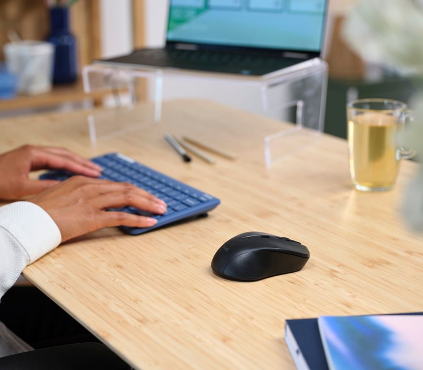 Imagen de un escritorio con una computadora portátil con gráficos en la pantalla, una taza de té, y manos tecleando en un teclado inalámbrico. Al lado del teclado hay un Mouse inalámbrico silencioso Trust Mydo de color negro