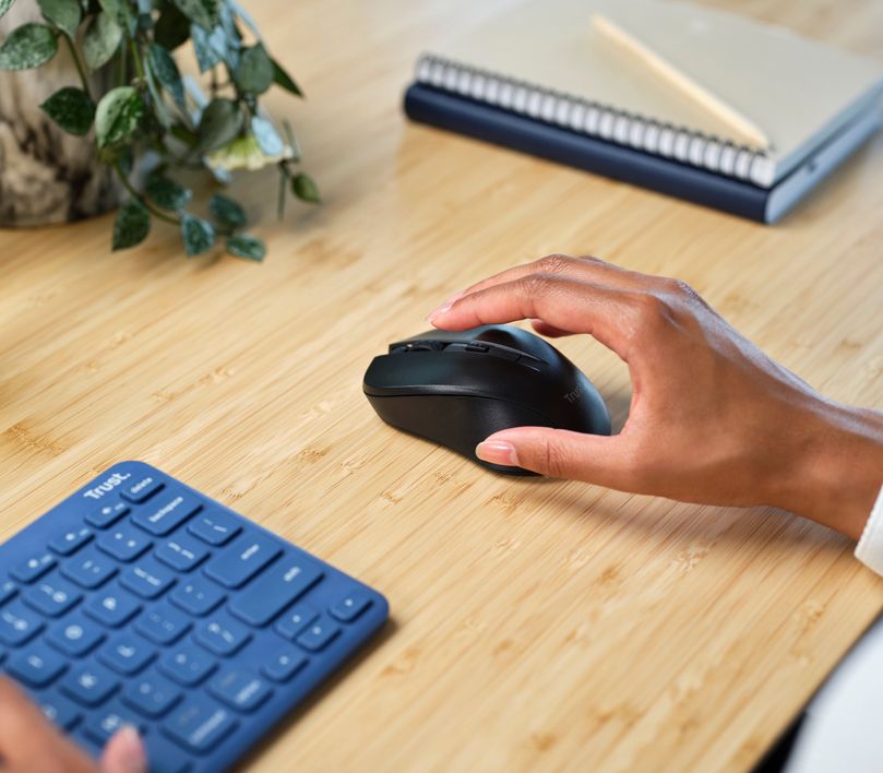 Una mano sobre un Mouse inalámbrico silencioso Trust Mydo de color negro al lado de un teclado y una libreta con un lápiz sobre una mesa de madera, con una planta parcialmente visible en el fondo.