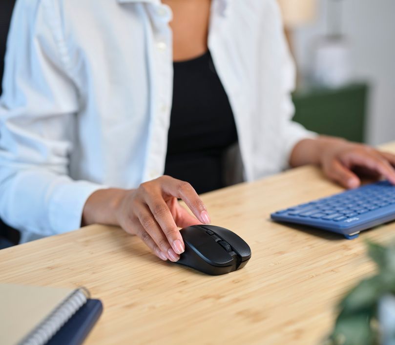 Una persona utiliza un Mouse inalámbrico silencioso Trust Mydo de color negro sobre un escritorio de madera, junto a un teclado y un cuaderno.