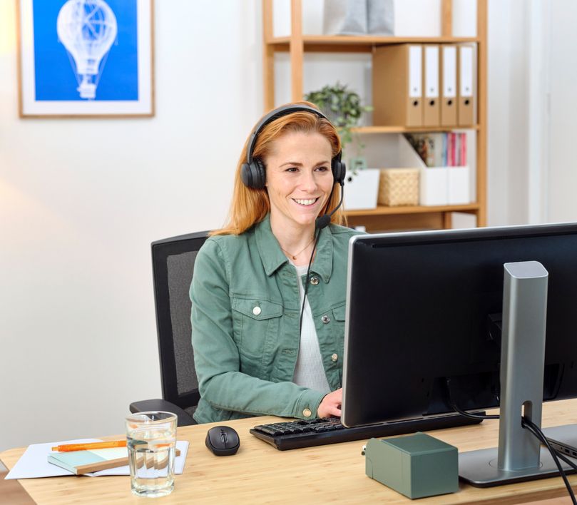 Una mujer con auriculares para PC Trust Ayda está sentada frente a una computadora en una oficina en casa. Tiene un vaso de agua y notas adhesivas sobre el escritorio, y hay una estantería al fondo.
