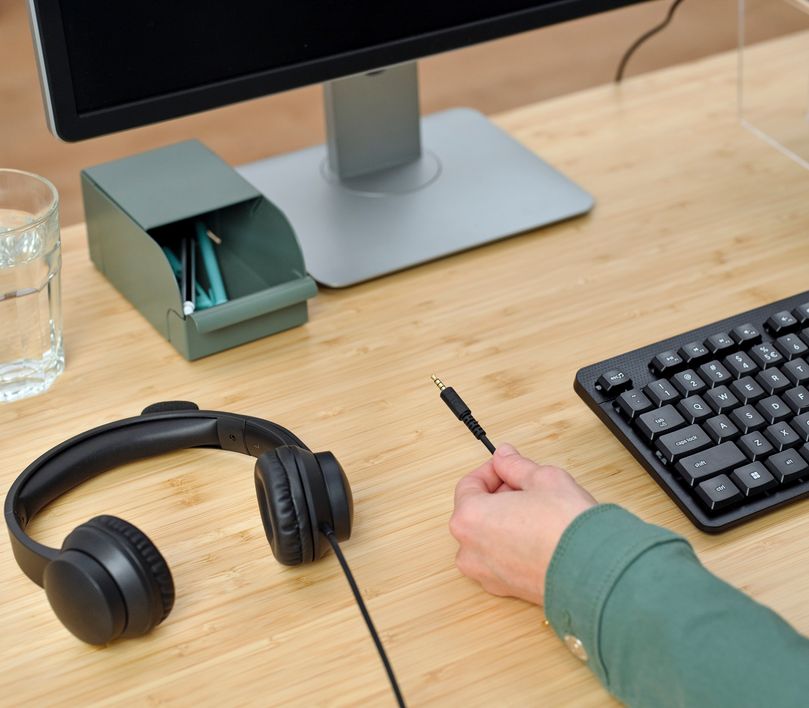Una mano sostiene el conector de audio de 3.5 mm de los auriculares para PC Trust Ayda cerca de una computadora de escritorio con monitor y un teclado sobre un escritorio de madera.