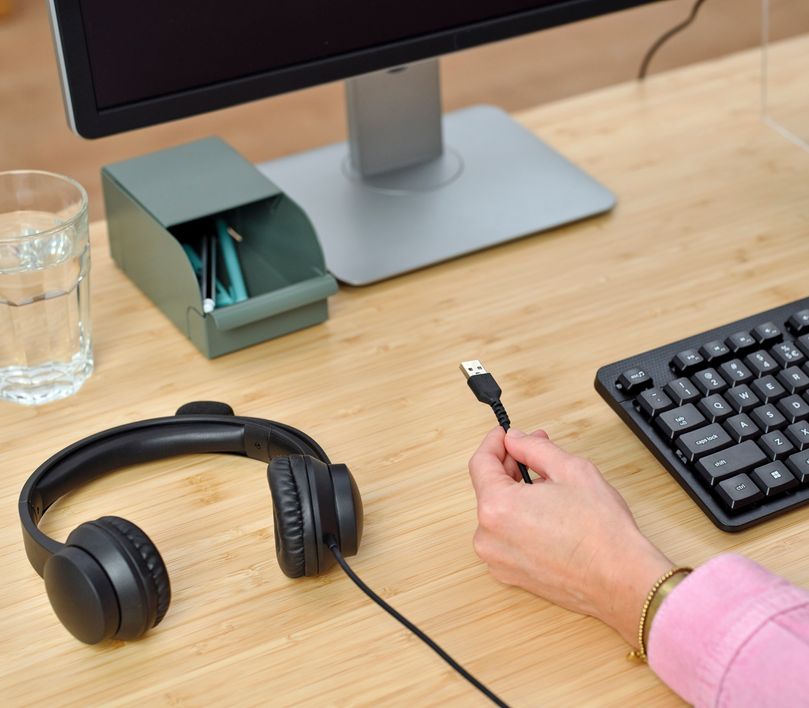 Una mano sostiene el conector USB de los auriculares USB para PC Trust Ayda cerca de una computadora de escritorio con monitor y un teclado sobre un escritorio de madera.