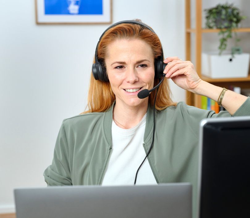 Una mujer con auriculares USB-ENC para PC Trust Ayda está sentada frente a una computadora en una oficina en casa. Hay una estantería al fondo.