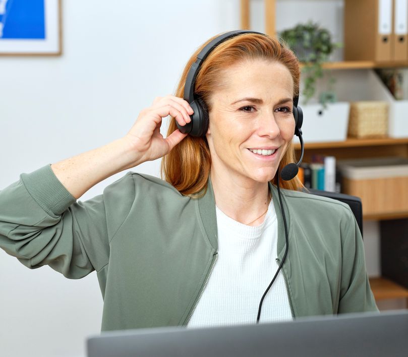 Una mujer con auriculares USB-ENC para PC Trust Ayda está sentada frente a una computadora en una oficina en casa.