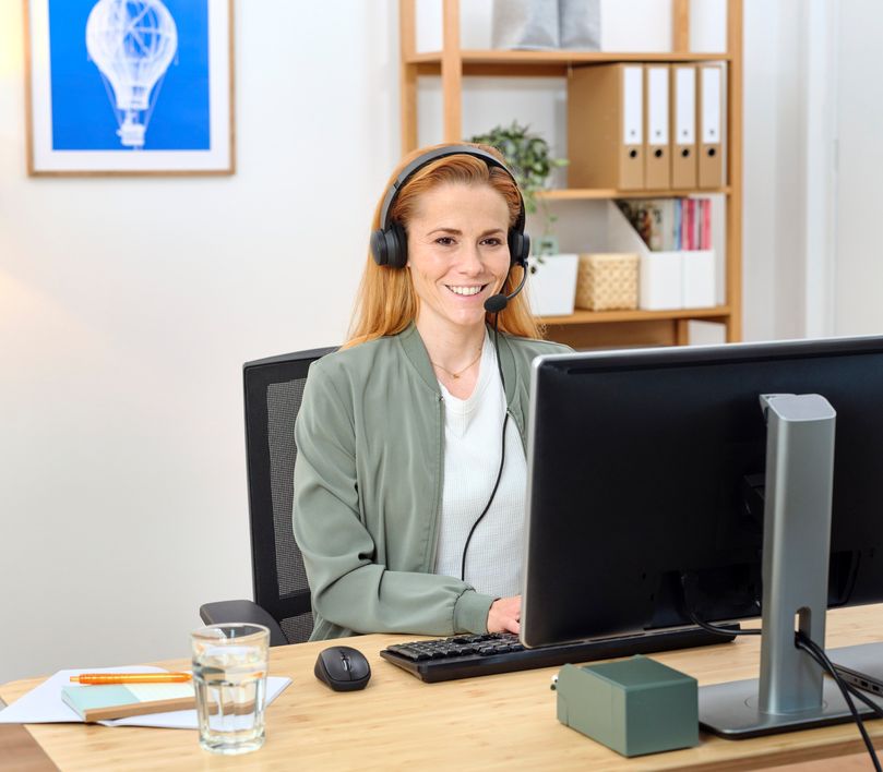 Persona con auriculares USB-ENC para PC Trust Ayda sentada frente a una computadora en un escritorio con un vaso de agua al lado, en una oficina con estantería.