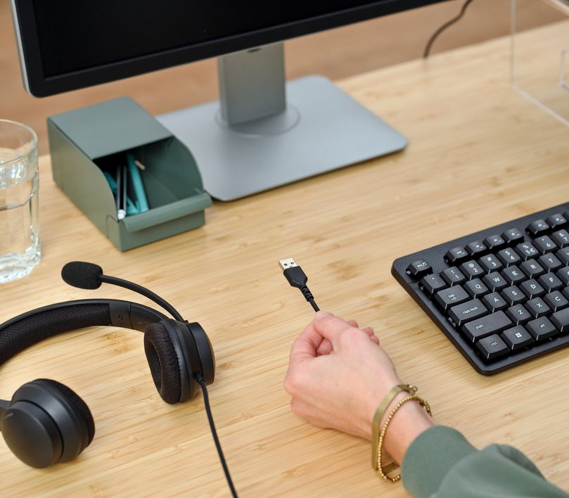 Una mano sostiene el conector USB de los auriculares USB-ENC para PC Trust Ayda cerca de una computadora de escritorio con monitor y un teclado sobre un escritorio de madera.