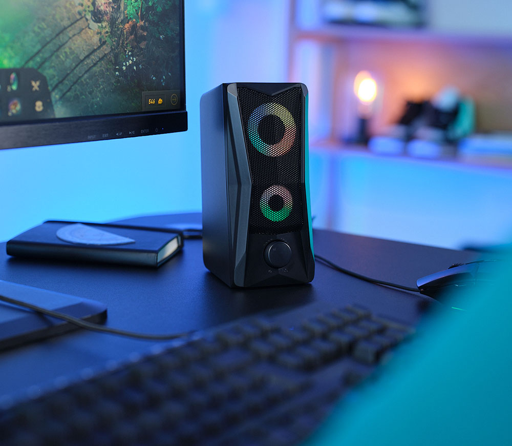 Parlante con iluminación RGB Trust GXT 606 Javv de color negro, sobre un escritorio con fondo desenfocado que incluye una pantalla de computadora y un cuaderno.