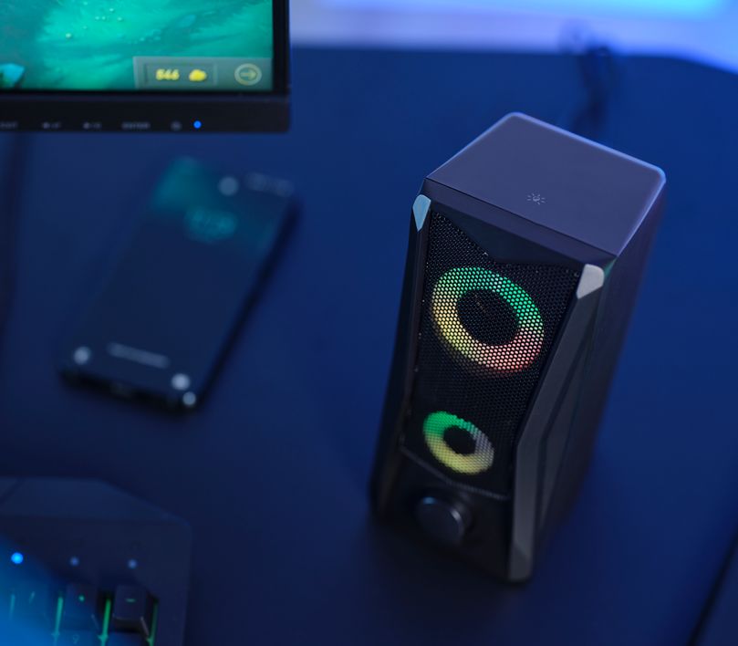 Parlante con iluminación RGB Trust GXT 606 Javv de color negro, en una habitación oscura, junto a un smartphone y una computadora.