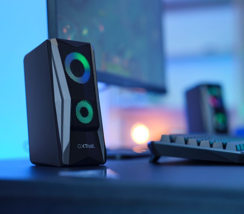 Parlante con iluminación RGB Trust GXT 606 Javv de color negro, situado sobre un escritorio con iluminación azulada y un teclado en el fondo desenfocado.